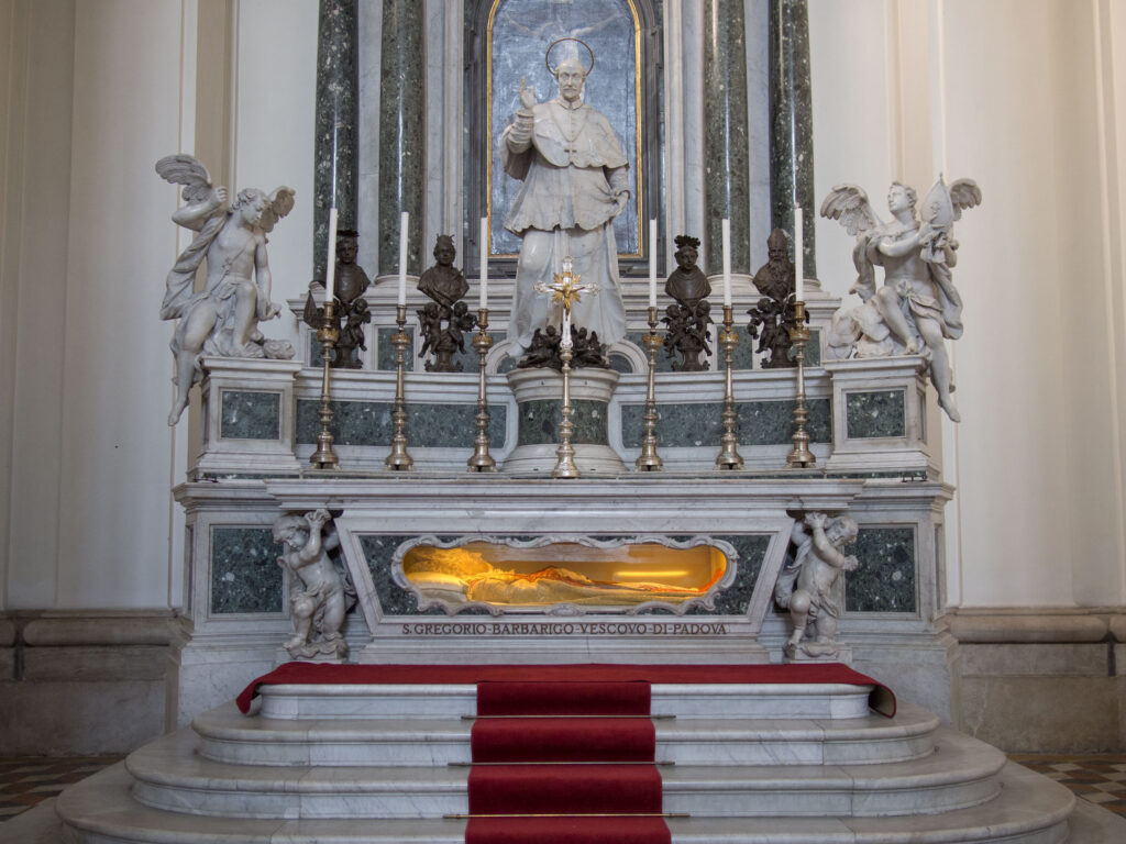 Photo of tomb of St. Gregorio Barbarigo. There is a window cut out of the wide of his coffin and the body, sheathed in robes, is illuminated with amber light.